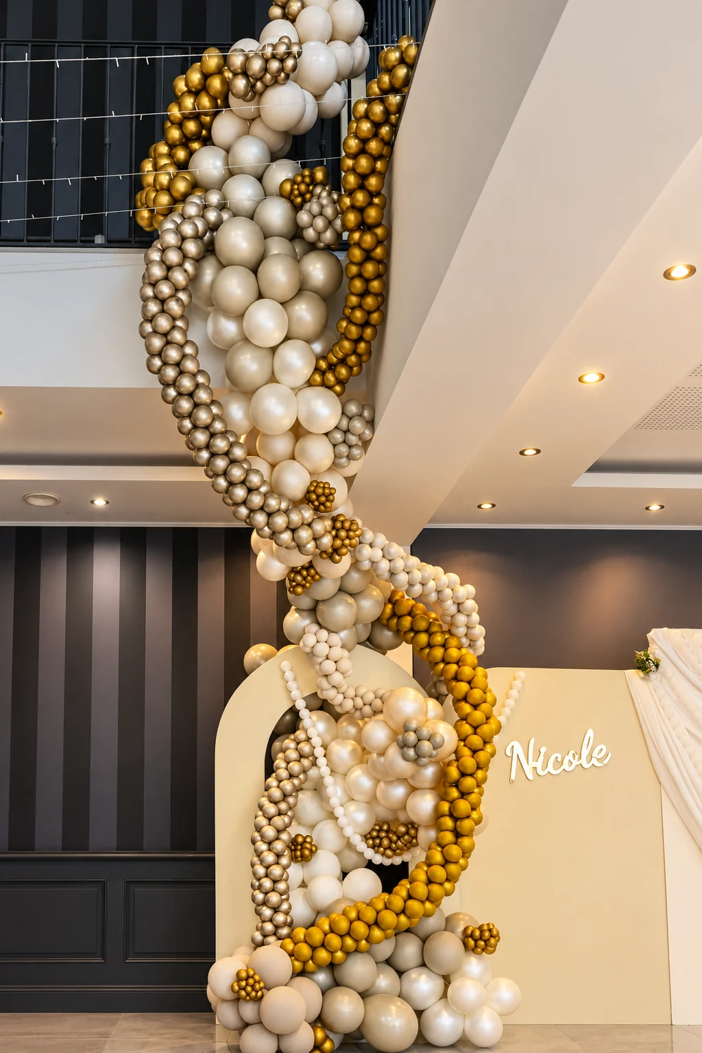 Cascade de ballons ivoire, nacrés et dorés descendant de la mezzanine devant le panneau Nicole